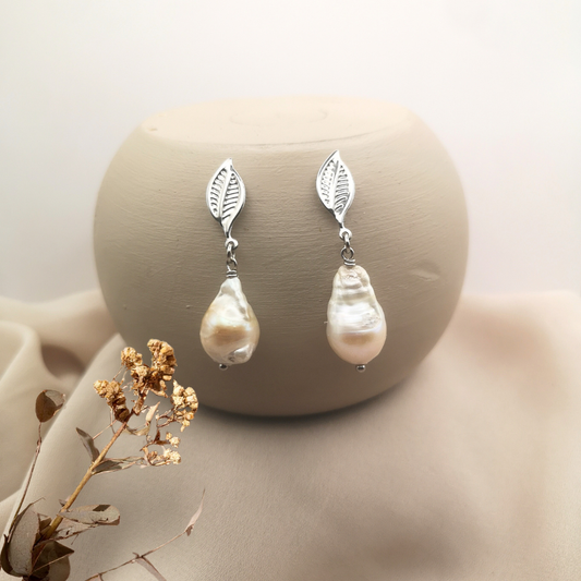 Stainless steel leaf stud earrings featuring unique white baroque freshwater pearls, displayed on a neutral beige background with dried floral accents.
