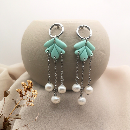 Handcrafted dangle earrings with aqua polymer clay petal shapes, stainless steel open ring studs, and three delicate chains adorned with white freshwater pearls, displayed on a neutral beige background with a soft dried flower accent.