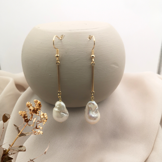 Gold-plated stainless steel dangle earrings with long bars and unique white baroque freshwater pearls, displayed on a soft beige background with dried floral accents.