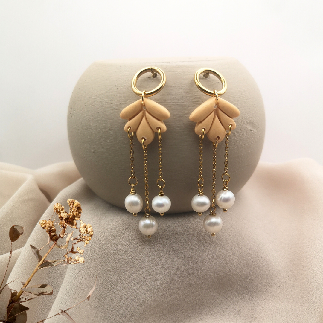 Handcrafted dangle earrings made of nude-toned polymer clay petals with gold-plated stainless-steel chains and white freshwater pearls, displayed on a soft beige fabric background.