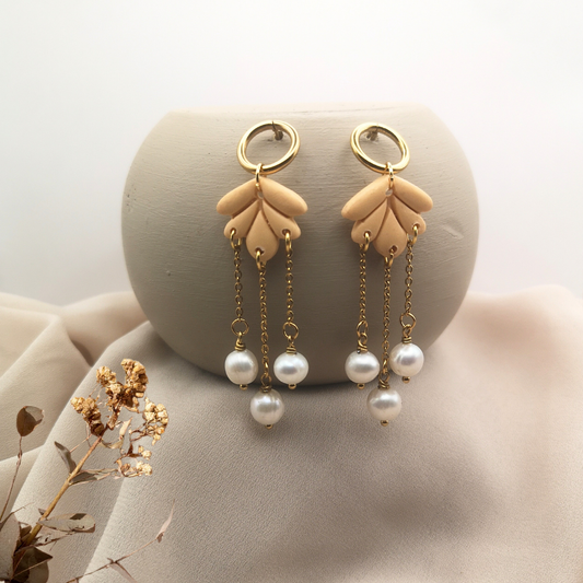 Handcrafted dangle earrings made of nude-toned polymer clay petals with gold-plated stainless-steel chains and white freshwater pearls, displayed on a soft beige fabric background.