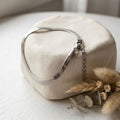 A silver snake chain necklace draped over a textured cream-colored stone display block, styled with dried grasses and eucalyptus on a white wooden surface.