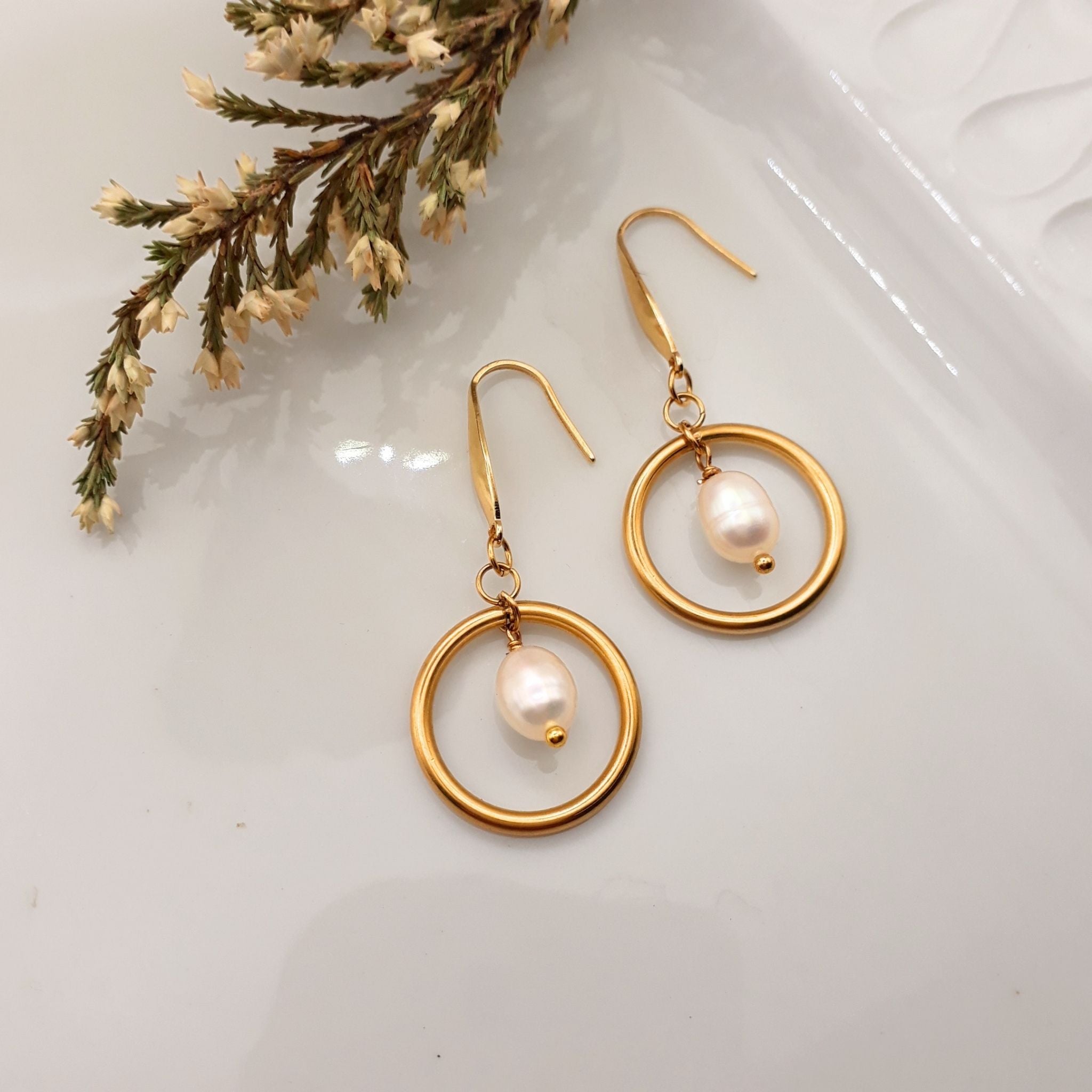 A pair of gold-tone drop earrings with open circular hoops and a small white pearl at the center, displayed on a glossy white surface with dried floral stems in the corner.
