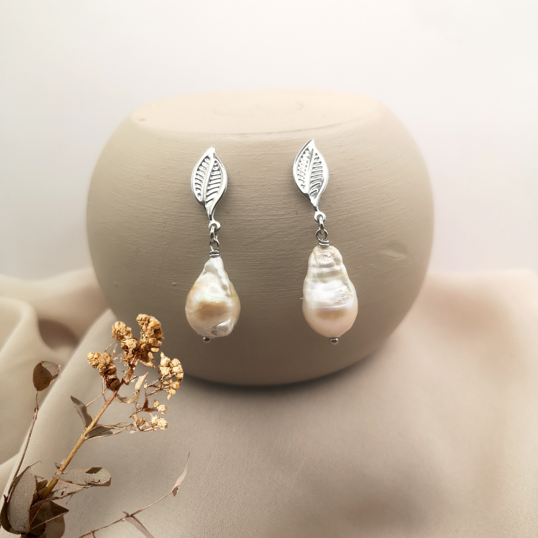 Stainless steel leaf stud earrings featuring unique white baroque freshwater pearls, displayed on a neutral beige background with dried floral accents.