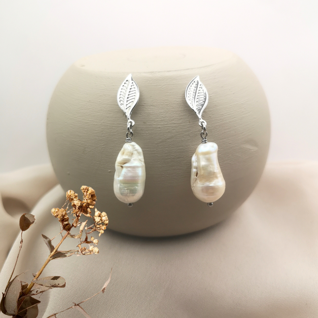 Stainless steel leaf stud earrings with unique white baroque freshwater pearls, displayed on a neutral beige background with dried floral accents for an elegant, natural look.