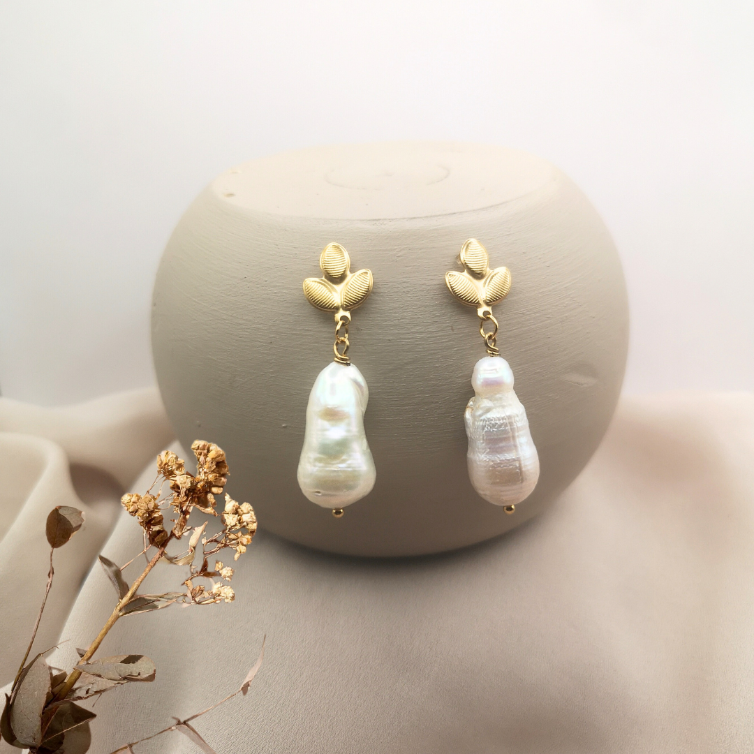 Gold-plated stainless steel leaf stud earrings featuring large white baroque freshwater pearls, displayed on a neutral beige background for an elegant and natural look.