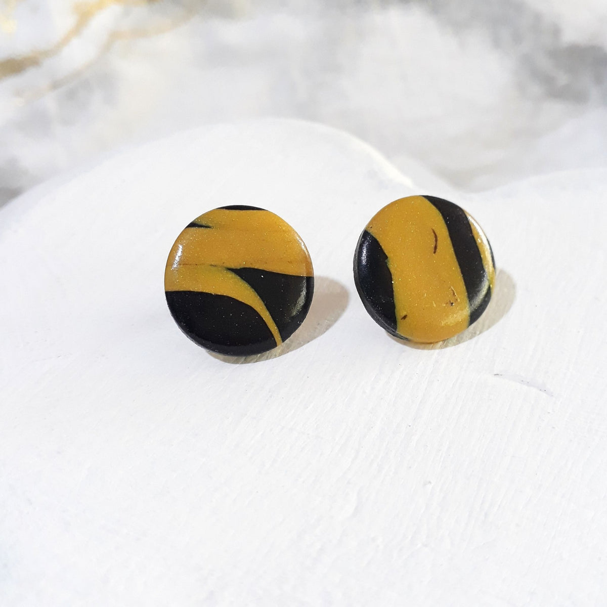 Glossy round polymer clay stud earrings with irregular black and mustard-yellow bands and curved colour patches.