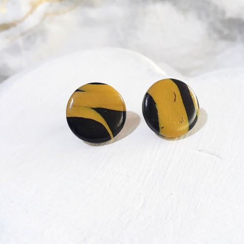 Glossy round polymer clay stud earrings with irregular black and mustard-yellow bands and curved colour patches.