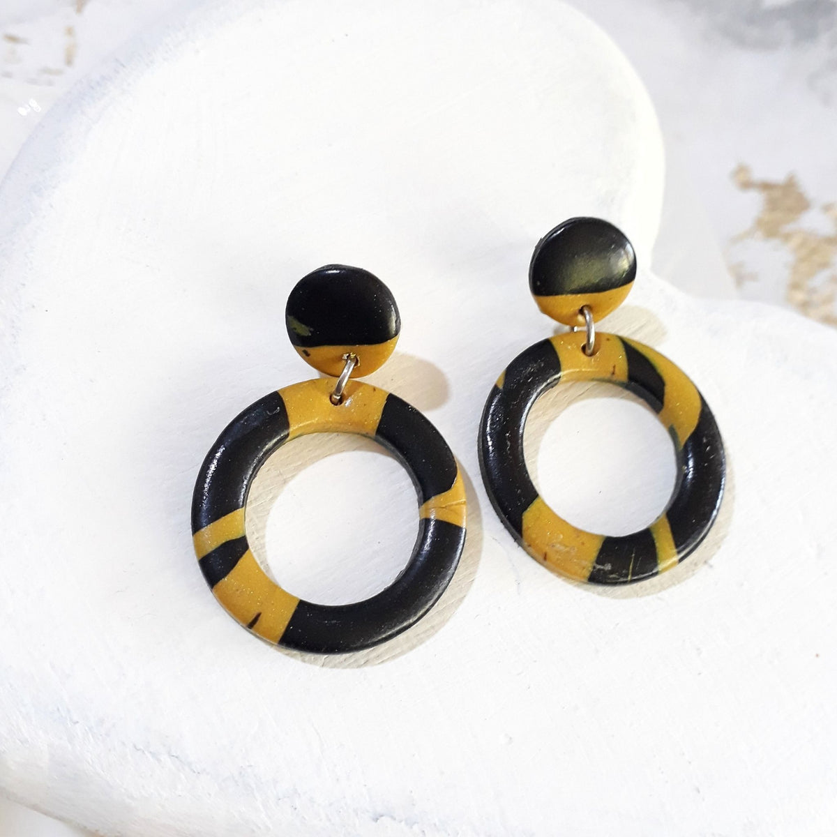 Glossy black and mustard-yellow polymer clay earrings with round stud tops and open-circle drops patterned with irregular contrasting bands.