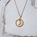 Gold-plated stainless steel necklace with a white freshwater pearl suspended inside an open circular pendant, displayed on a textured white surface.

