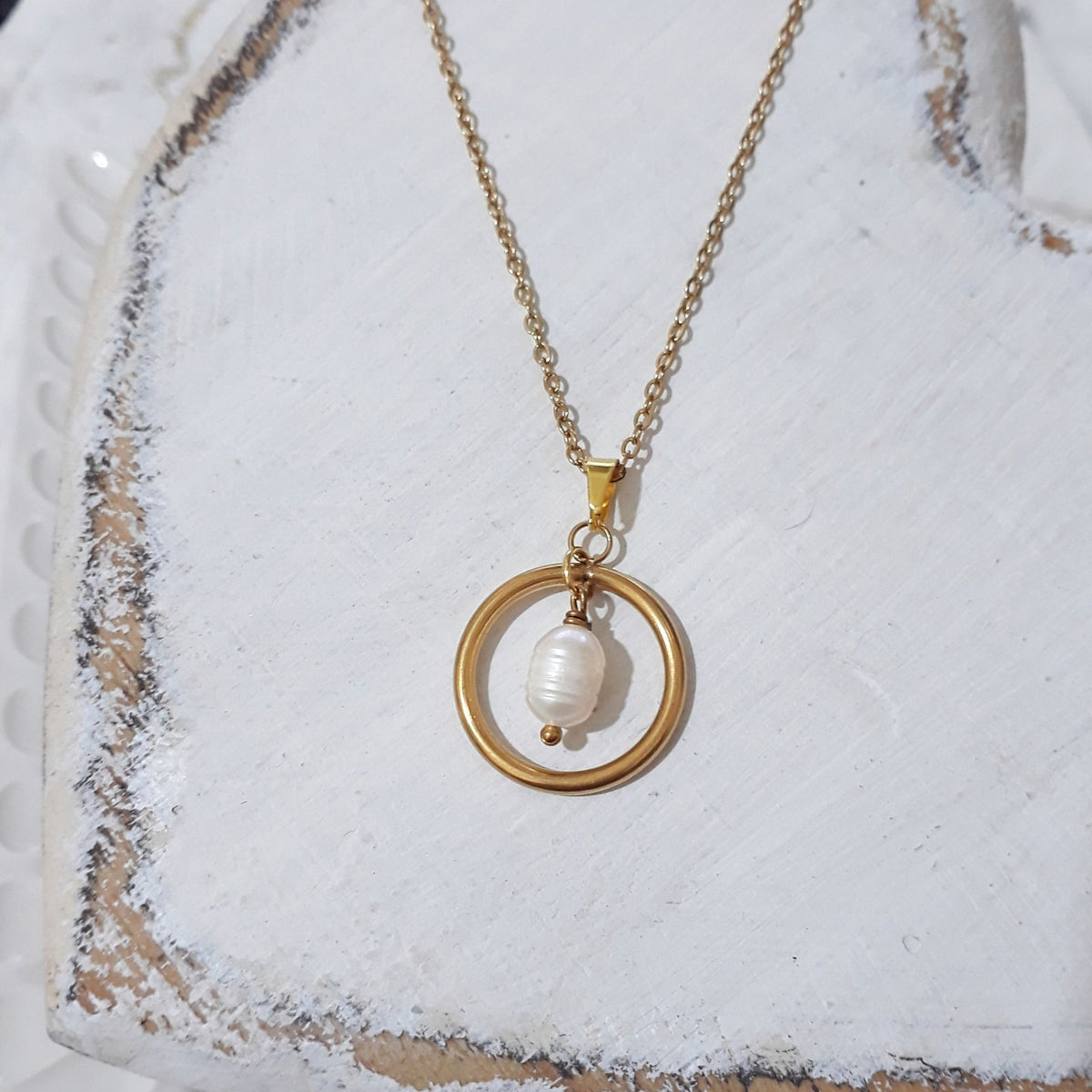 Gold-plated stainless steel necklace with a white freshwater pearl suspended inside an open circular pendant, displayed on a textured white surface.

