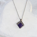 Diamond-shaped stainless steel pendant with purple crushed amethyst sealed in resin and a textured upper border on a silver-tone chain.