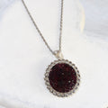 Round deep red crushed garnet pendant surrounded by clear rhinestones on a silver-tone stainless steel chain.
