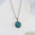 Round crushed turquoise pendant with blue-green pieces and dark markings in a silver-tone stainless steel bezel on a chain.