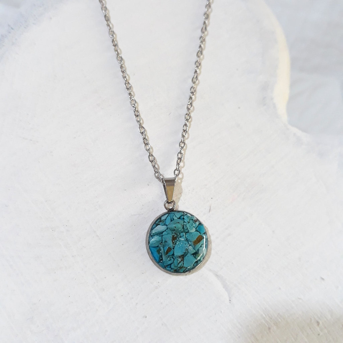 Round crushed turquoise pendant with blue-green pieces and dark markings in a silver-tone stainless steel bezel on a chain.