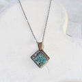 Diamond-shaped stainless steel pendant with blue-green crushed turquoise sealed in resin and a textured upper border on a silver-tone chain.