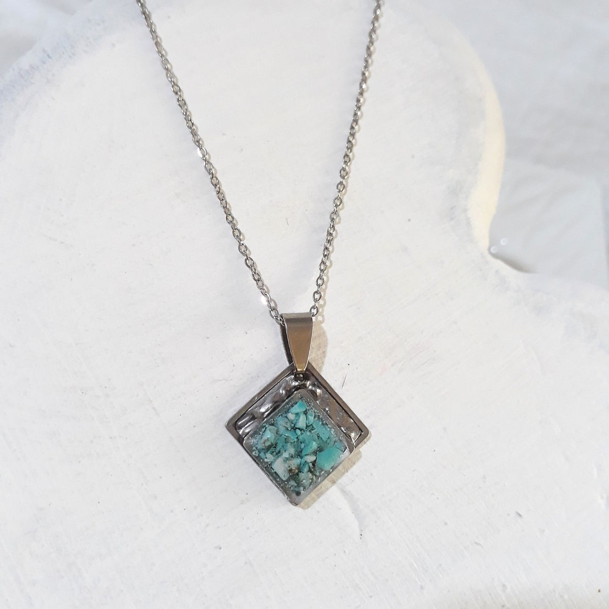 Diamond-shaped stainless steel pendant with blue-green crushed turquoise sealed in resin and a textured upper border on a silver-tone chain.