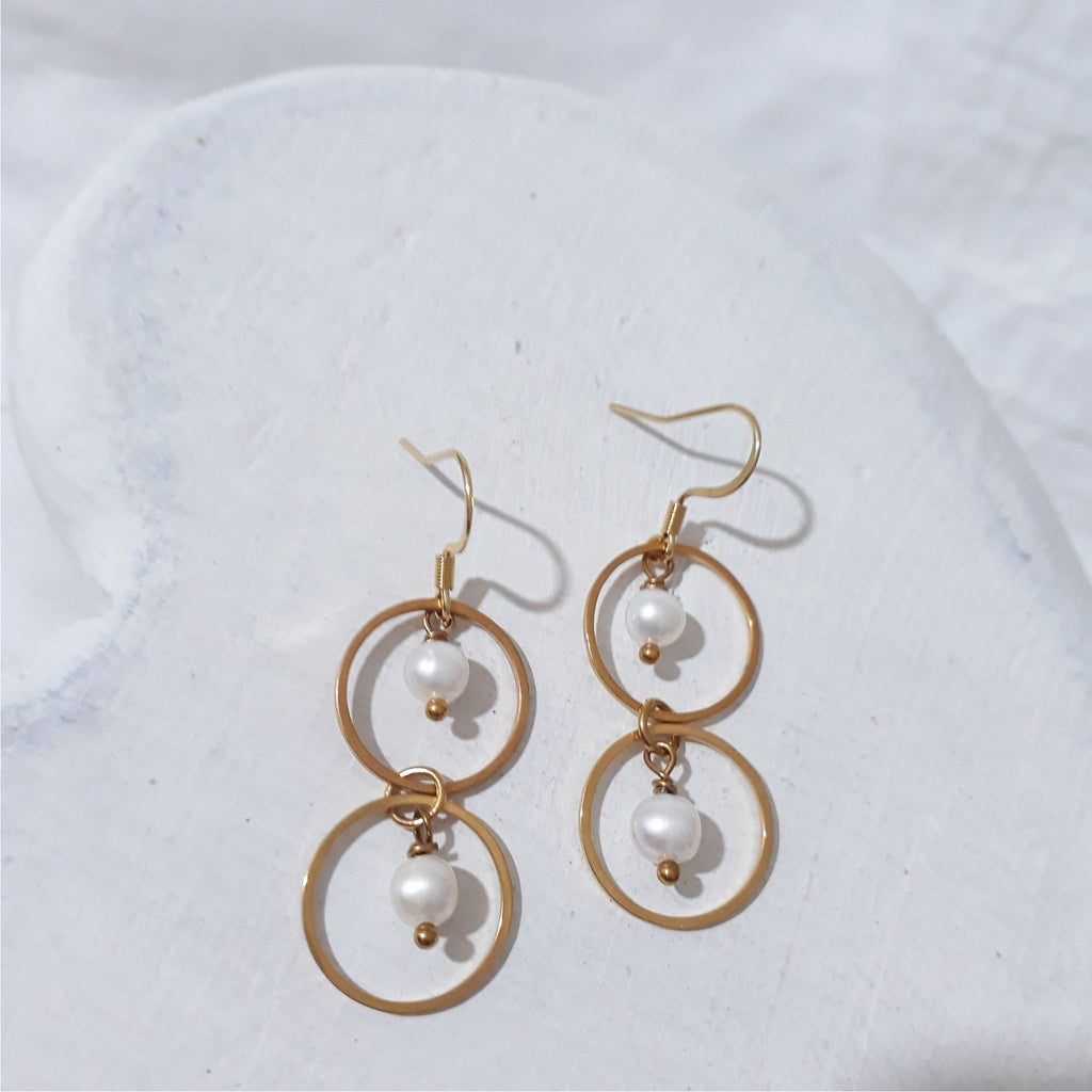 Gold-plated stainless steel double circle dangle earrings with a white freshwater pearl suspended inside each circle and shepherd hooks.