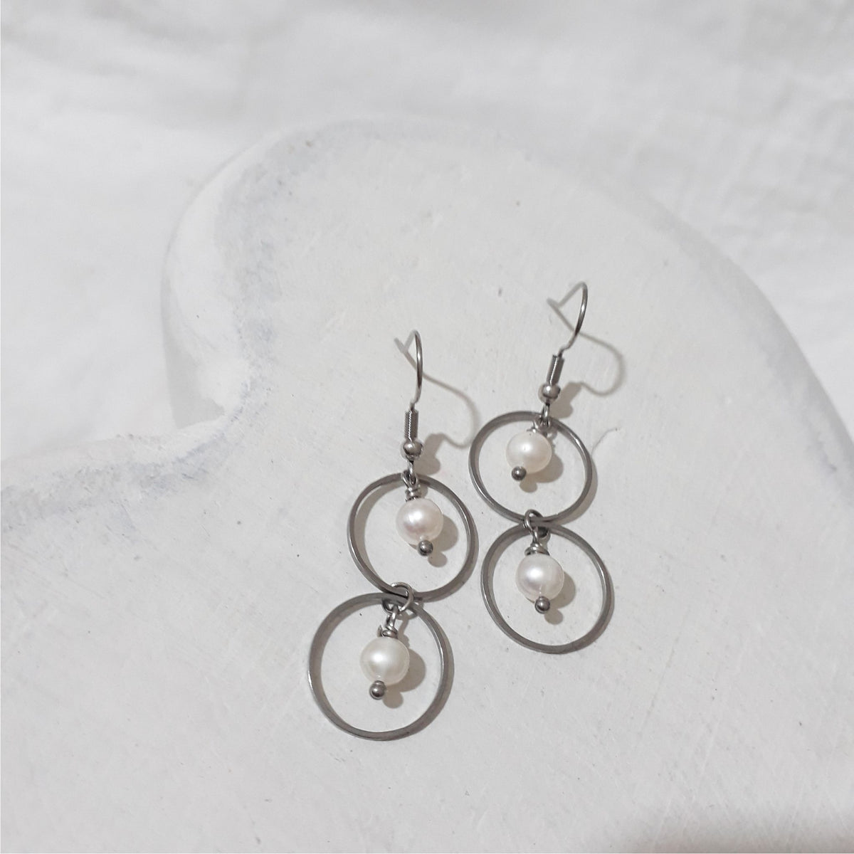 Stainless steel double circle dangle earrings with a white freshwater pearl suspended inside each circle and shepherd hooks.