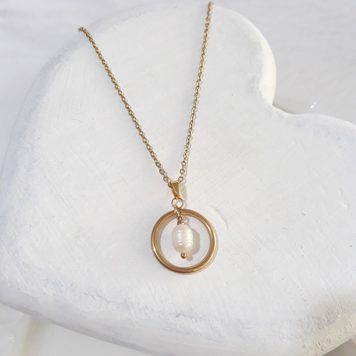 Gold-plated stainless steel necklace with a white freshwater pearl suspended inside an open circular pendant, displayed on a textured white surface.
