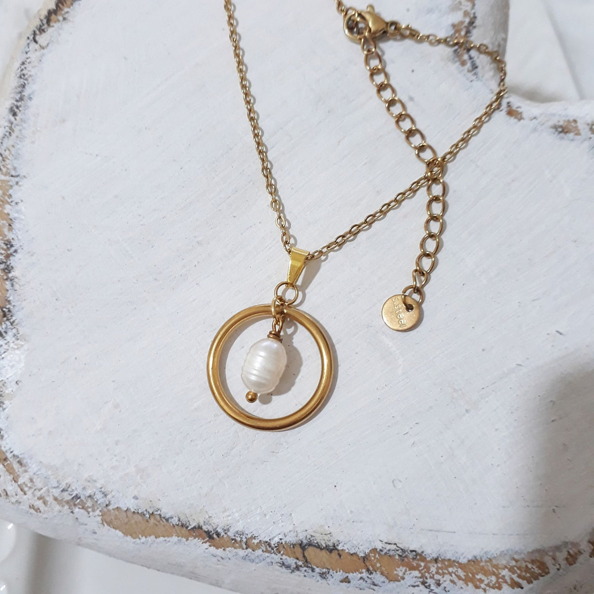A gold chain necklace with a circular open ring pendant enclosing a white baroque pearl, displayed on a distressed white wooden surface.