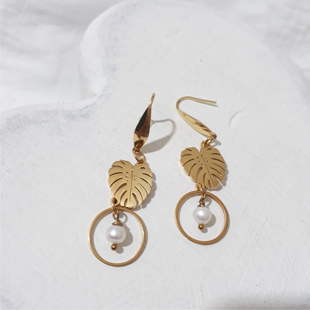 Gold-plated monstera leaf dangle earrings with round white freshwater pearls suspended inside open circles beneath the leaves.