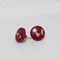 A pair of round, deep red polymer clay stud earrings with scattered gold leaf flakes, displayed on a white textured ceramic plate.