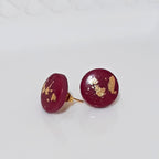 A pair of round, deep red polymer clay stud earrings with scattered gold leaf flakes, displayed on a white textured ceramic plate.