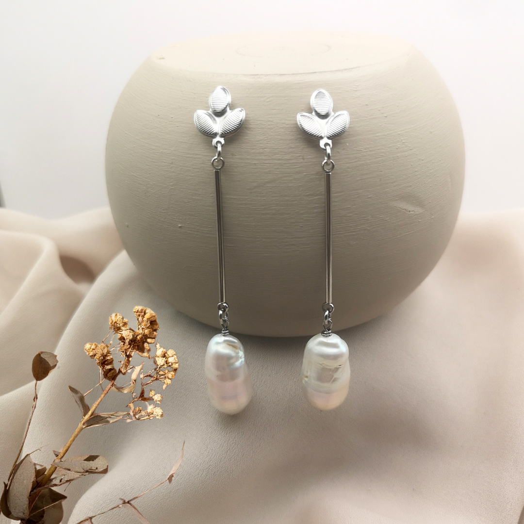 Stainless steel leaf stud earrings with long 30 mm bars and large white baroque freshwater pearls, displayed on a neutral beige background with dried floral accents.