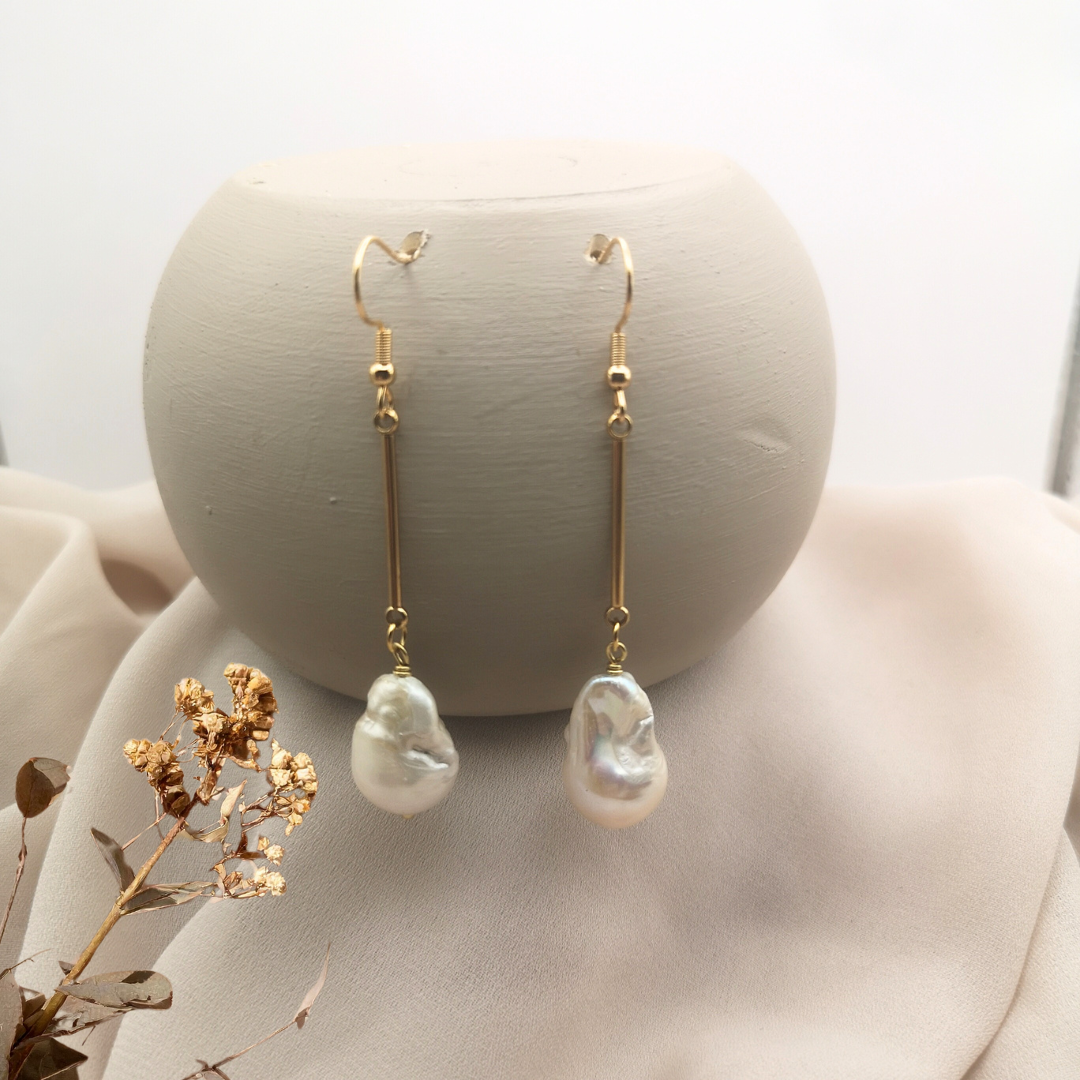 Gold-plated stainless steel dangle earrings with long bars and unique white baroque freshwater pearls, displayed on a soft beige background with dried floral accents.