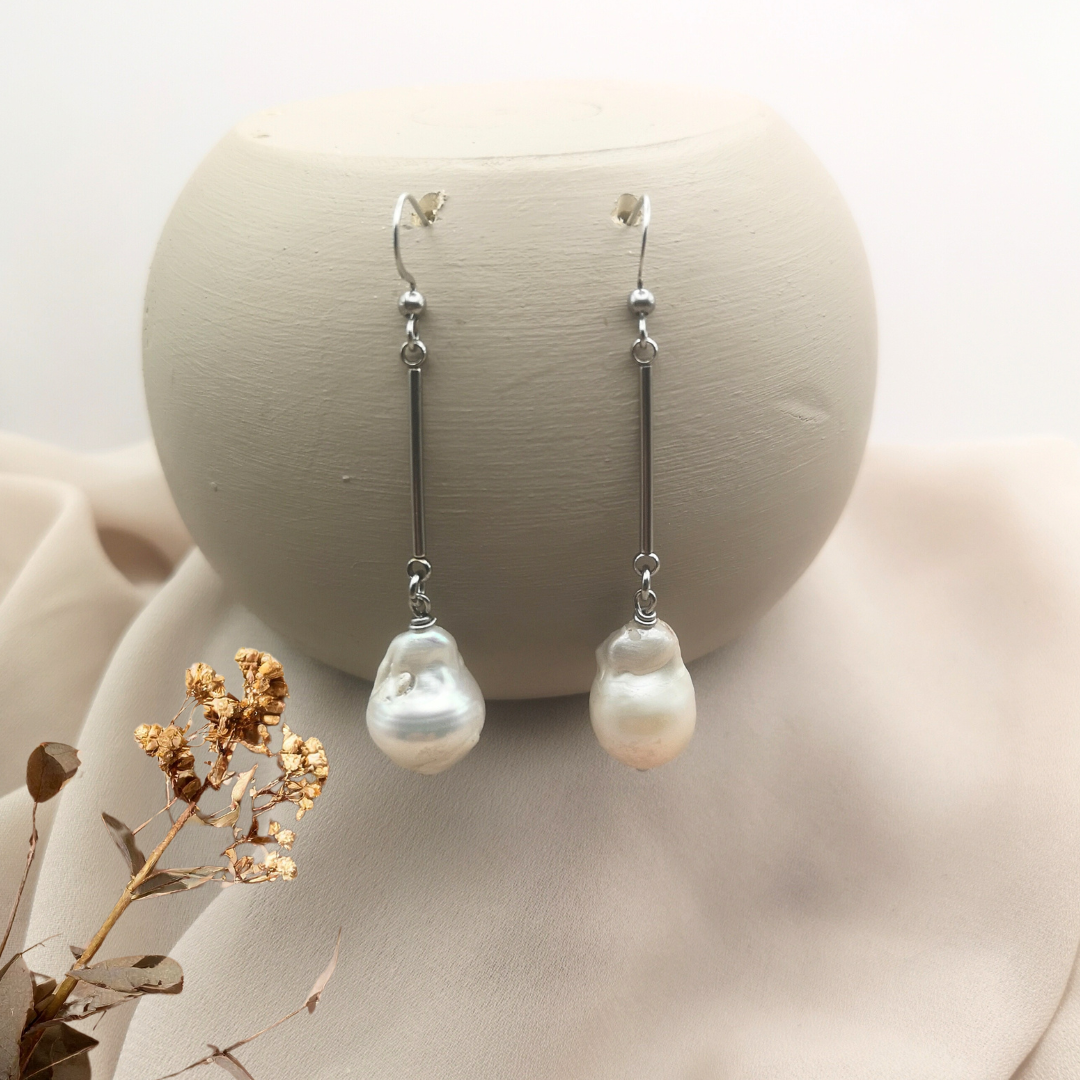 Stainless steel shepherd hook earrings with 20 mm bar drops and unique white baroque freshwater pearls, displayed on a neutral beige fabric background with dried floral accents.