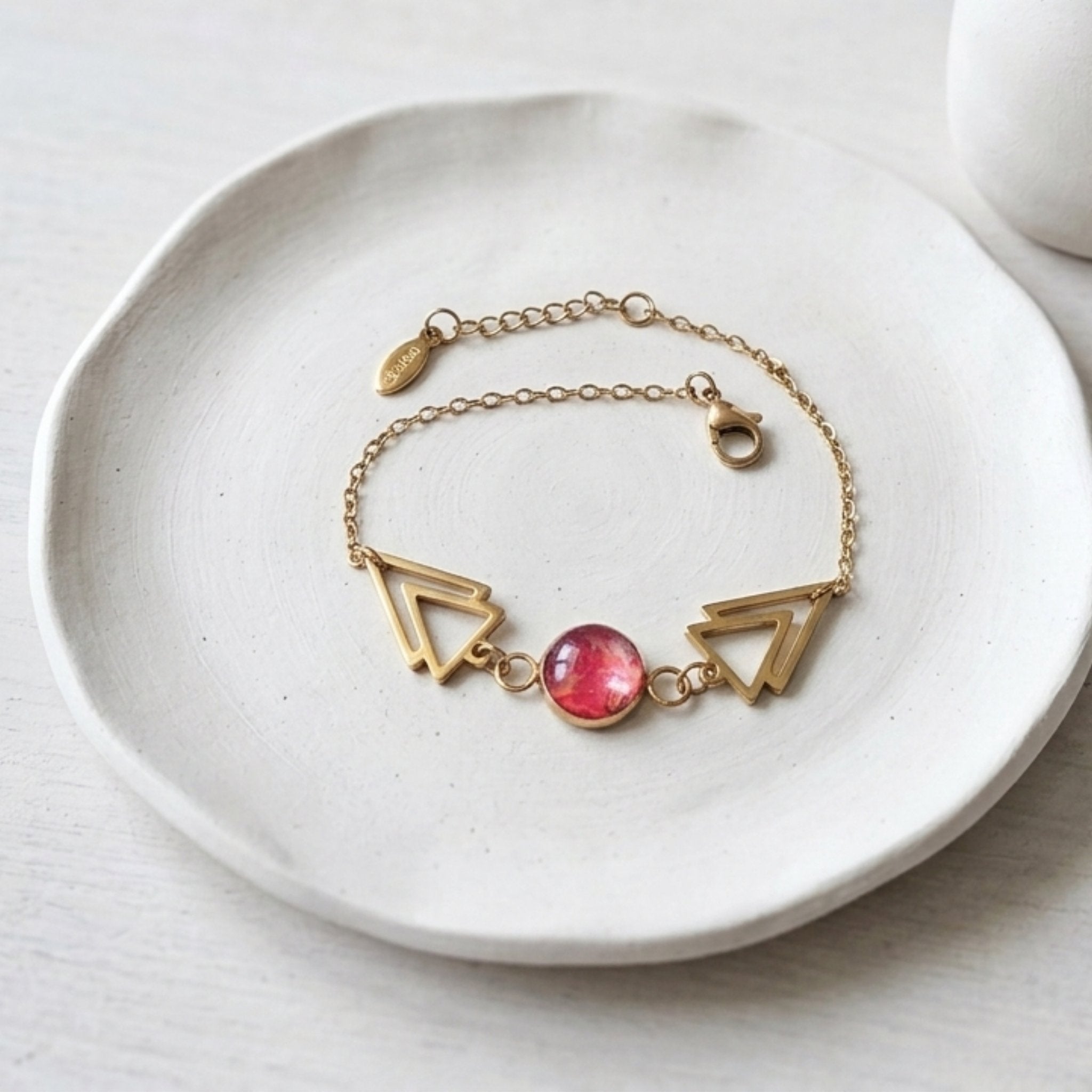 A delicate gold chain bracelet with two open triangle charms and a round pink gemstone centerpiece, displayed on a white ceramic dish.