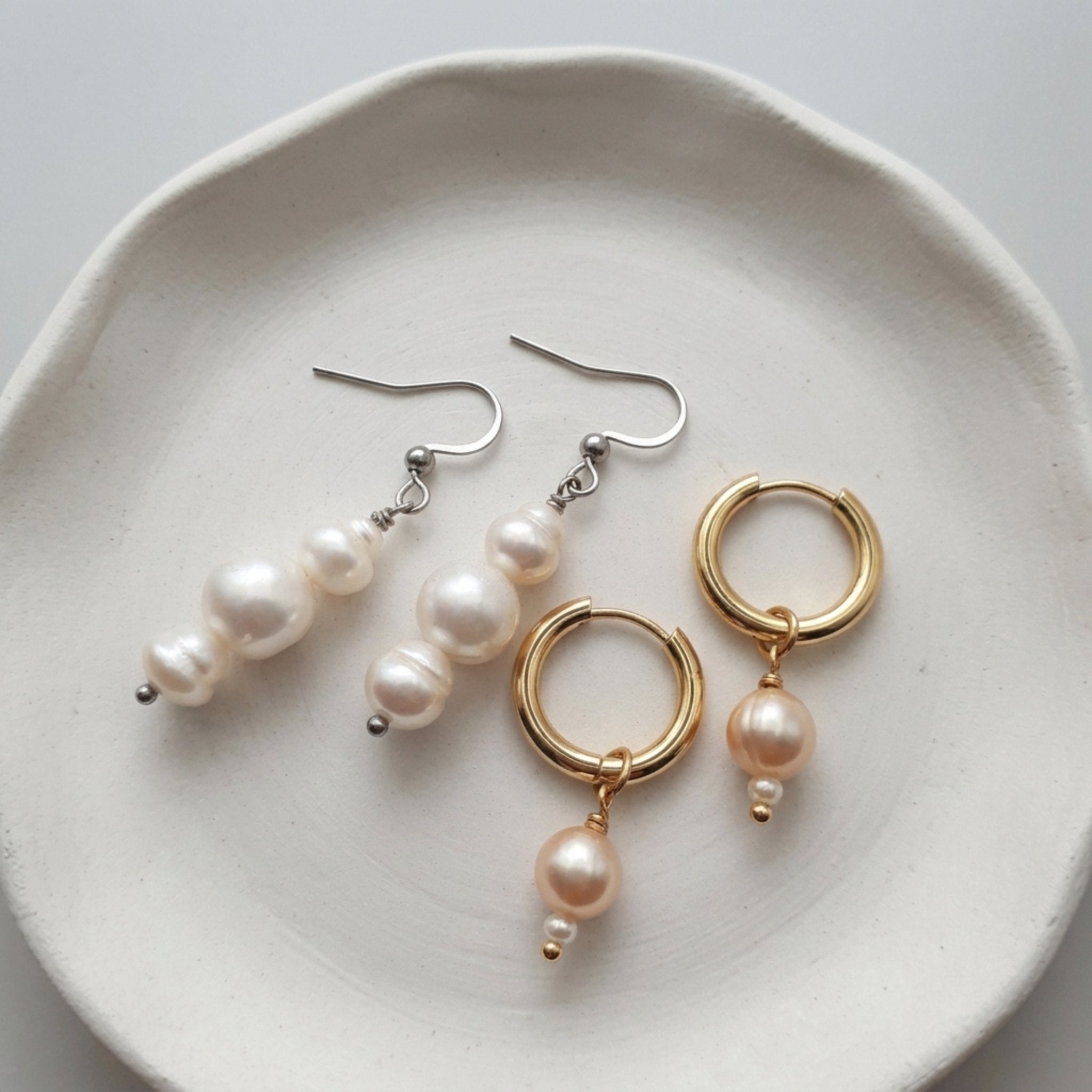 A pair of gold hoop earrings with dangling pearl drops and a pair of silver fishhook earrings with larger pearl clusters, displayed on a textured white ceramic plate.