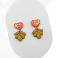 Heart-shaped orange earrings with gold accents and green leaf-like drops on a white background
