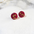 A pair of round, deep red polymer clay stud earrings with scattered gold leaf flakes, displayed on a white textured ceramic plate.