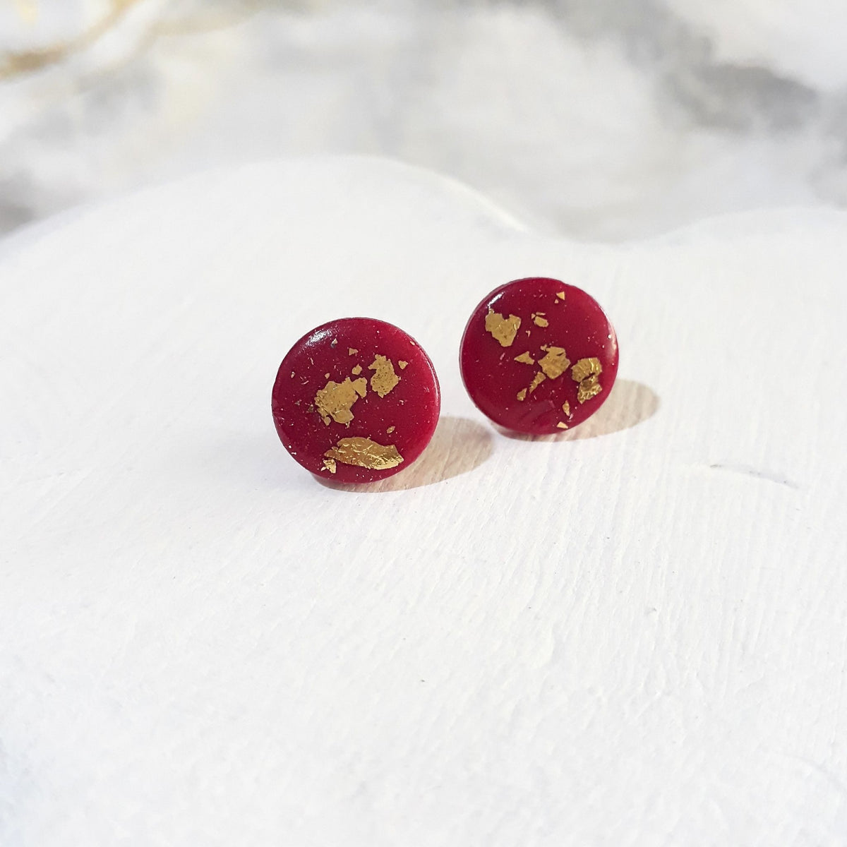 A pair of round, deep red polymer clay stud earrings with scattered gold leaf flakes, displayed on a white textured ceramic plate.