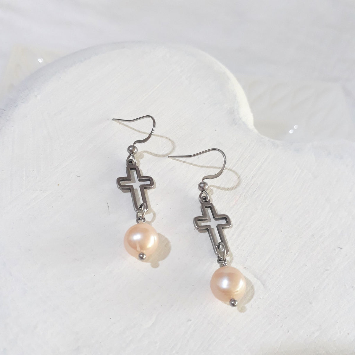 Soft peach freshwater pearls suspended beneath open silver-tone crosses on stainless steel ear hooks.