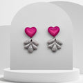 Pair of earrings with pink heart-shaped tops and silver leaf-like bottoms on a white stand.