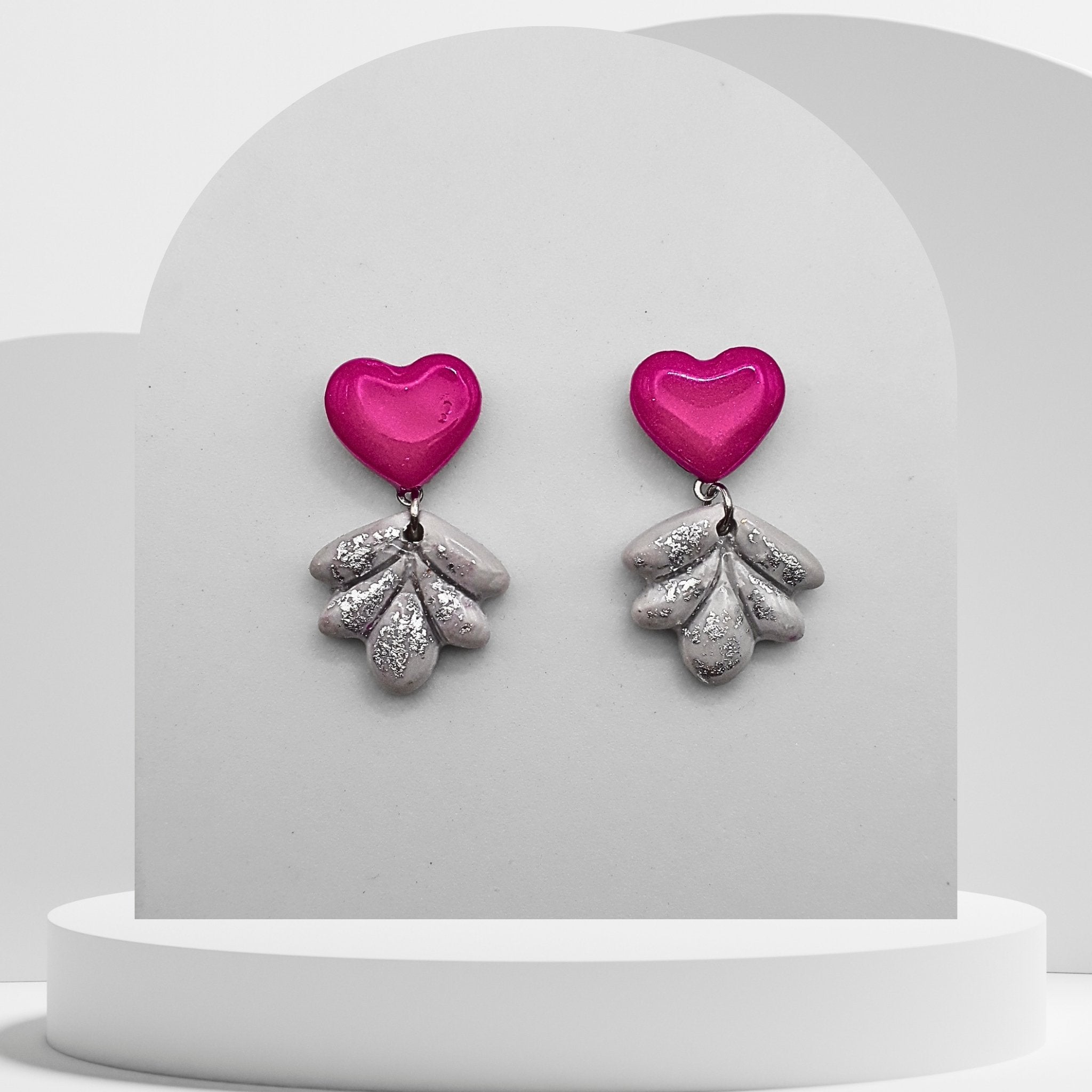 Pair of earrings with pink heart-shaped tops and silver leaf-like bottoms on a white stand.