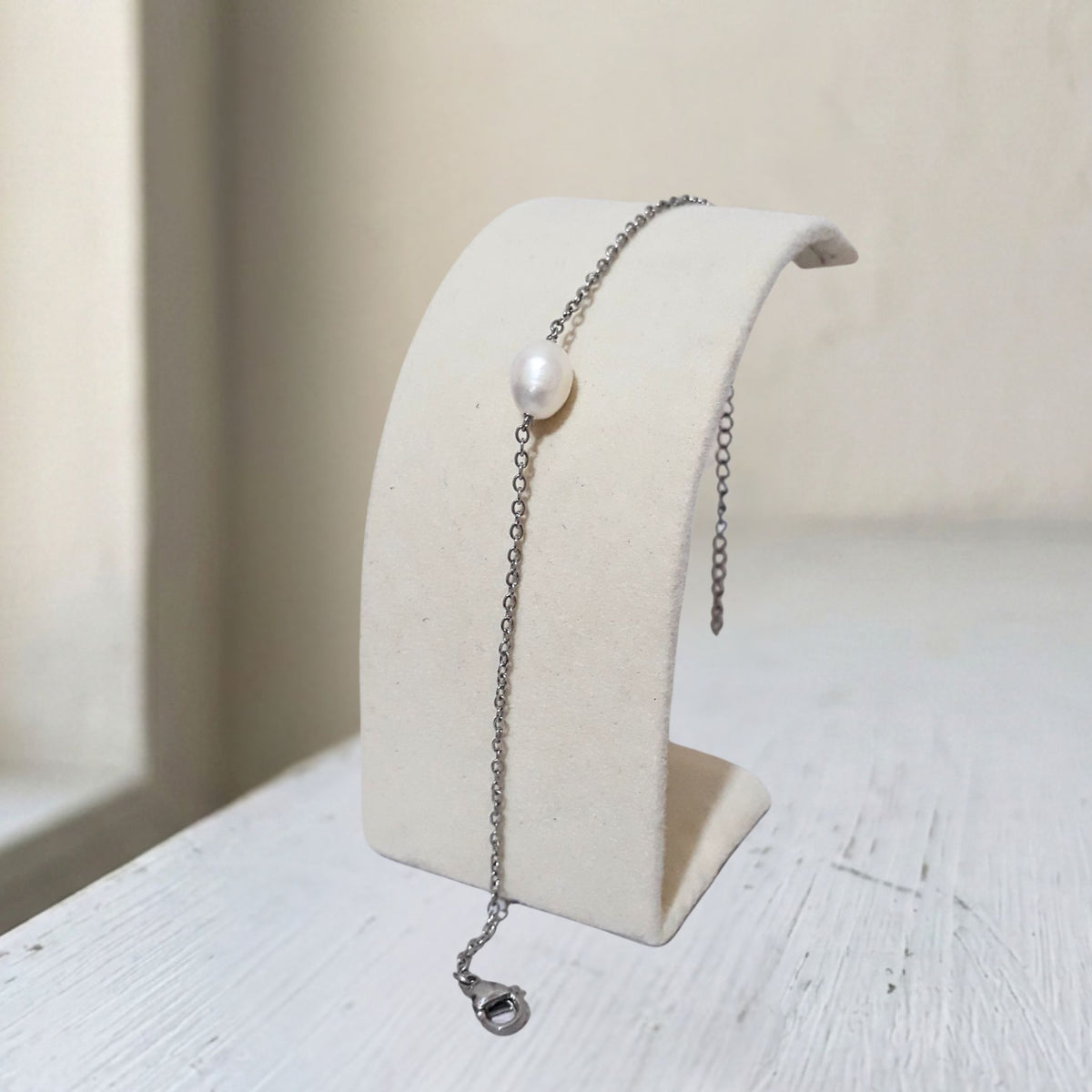 A delicate silver chain bracelet with a single white pearl, displayed on a cream-colored jewelry stand.