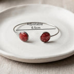 An adjustable silver-tone cuff bracelet with two round red stone cabochons, displayed on a textured white ceramic plate with a black measurement line and text indicating 'Adjustable 6.5cm'.