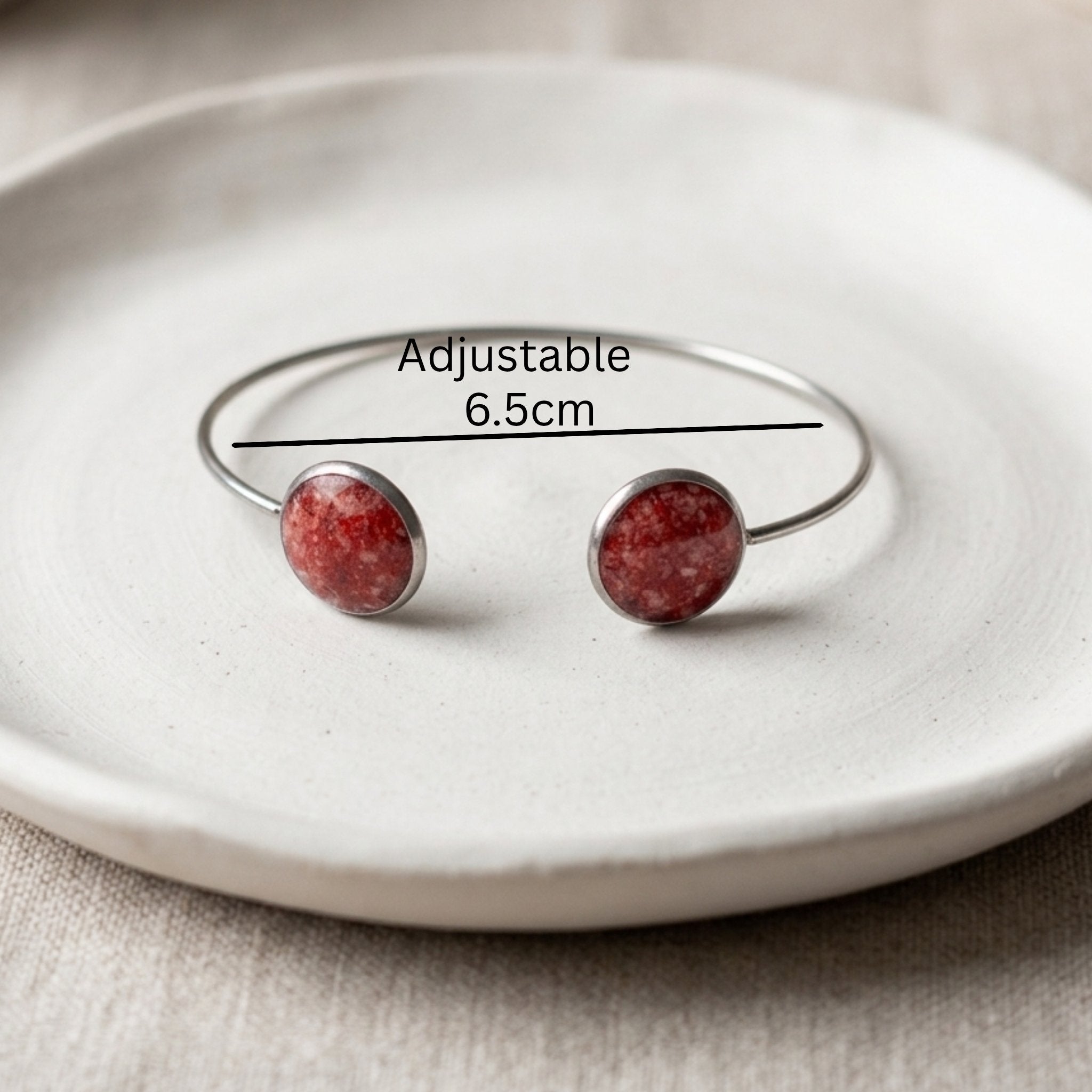 An adjustable silver-tone cuff bracelet with two round red stone cabochons, displayed on a textured white ceramic plate with a black measurement line and text indicating 'Adjustable 6.5cm'.