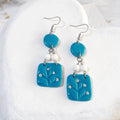 Turquoise polymer clay earrings with round upper pieces, white stone beads and rounded square botanical drops on silver-tone shepherd hooks.