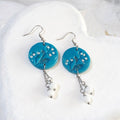 Turquoise round polymer clay earrings with leafy detailing, small white accents and white bead drops on silver-tone chains beneath shepherd hooks.