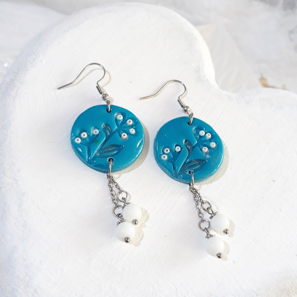 Turquoise round polymer clay earrings with leafy detailing, small white accents and white bead drops on silver-tone chains beneath shepherd hooks.