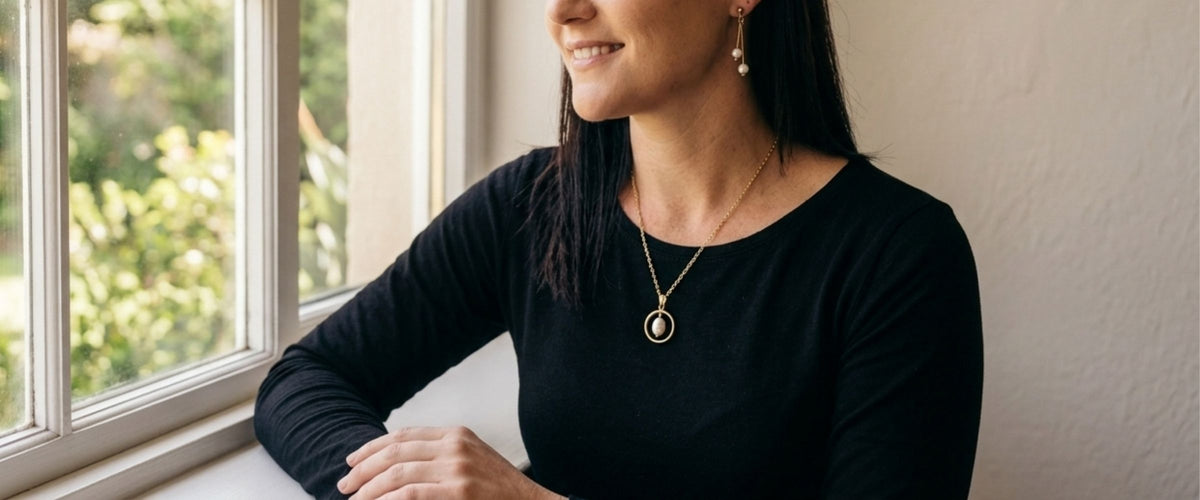 A woman with dark hair, wearing a black long-sleeve top and a gold oval pendant necklace, sitting by a window.