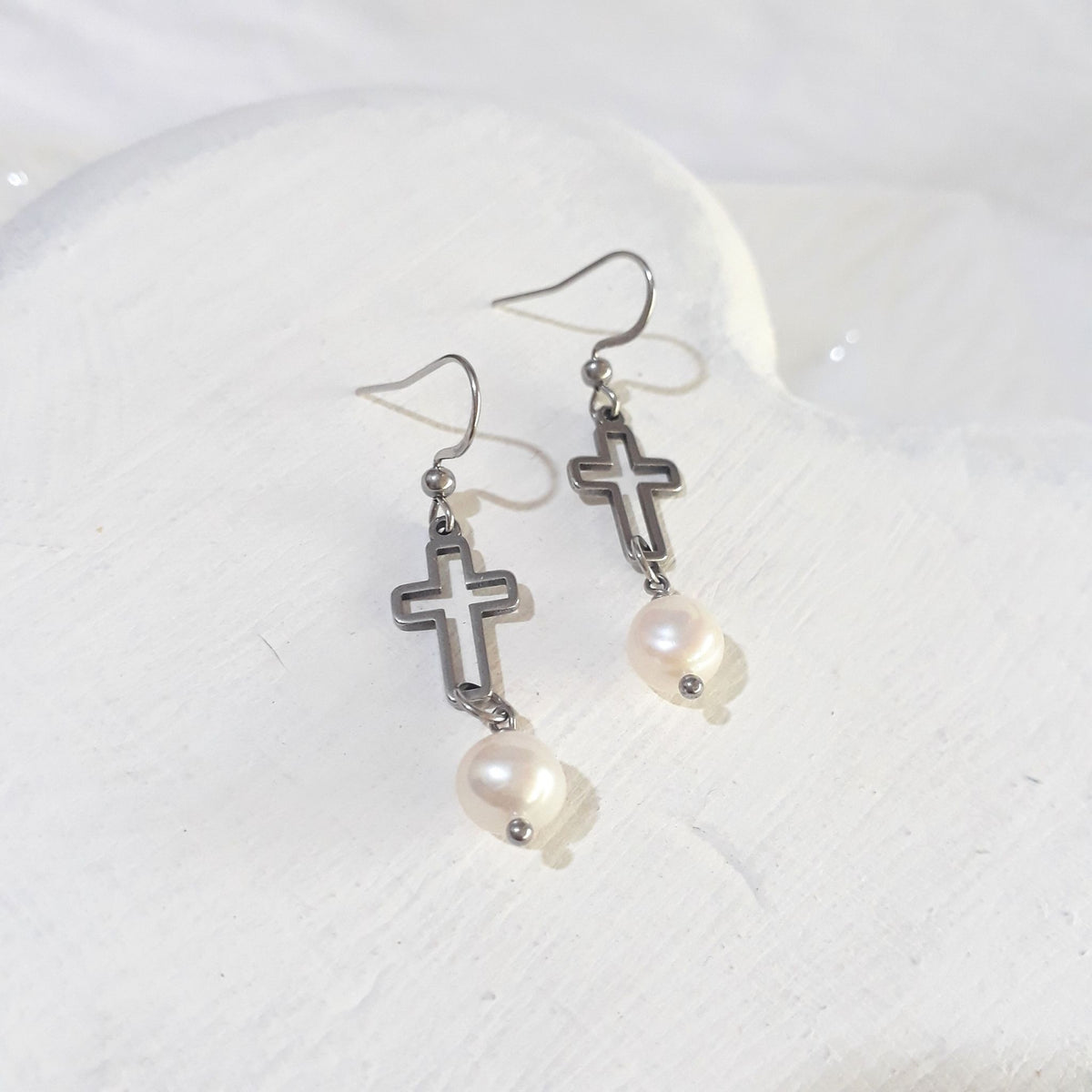 White freshwater pearls hanging beneath open silver-tone crosses on ear hooks, displayed on a white background.