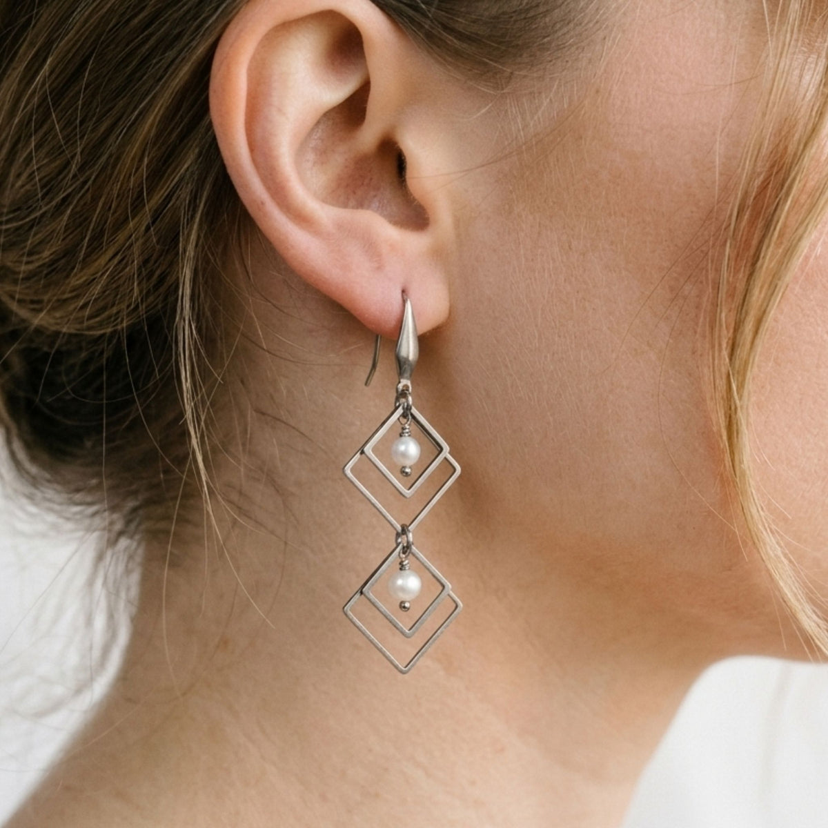 Close-up of a woman wearing a long stainless steel linked diamond-shaped dangle earring with two white freshwater pearls.
