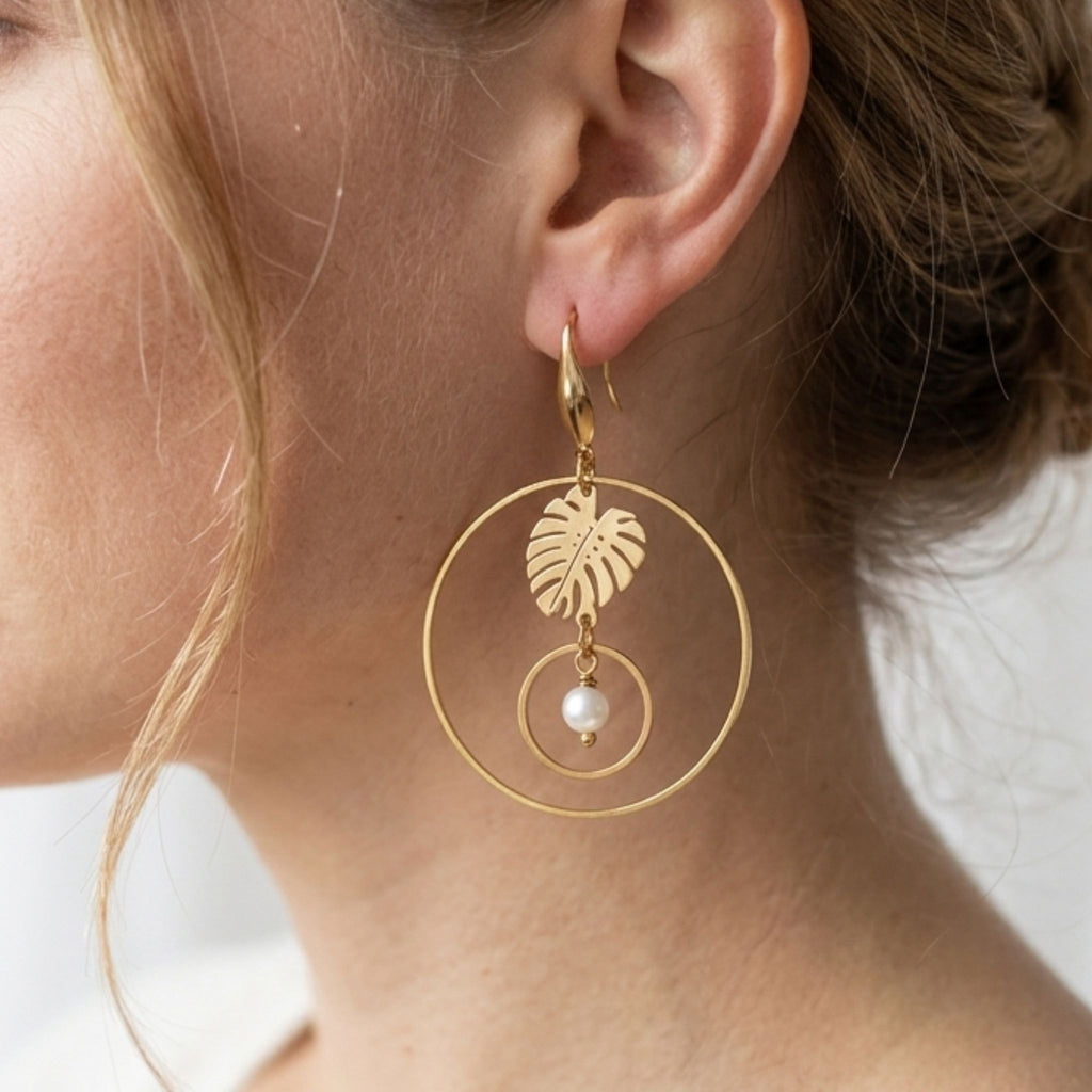 Woman wearing a gold-plated hoop earring with a monstera leaf and a white freshwater pearl suspended inside a smaller circle.