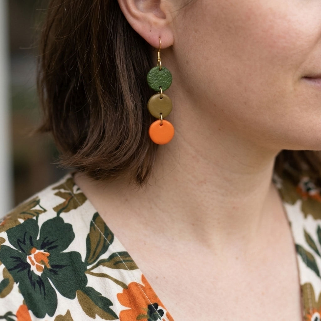 Woman wearing moss green, olive and orange three-circle polymer clay cascade earrings with gold-plated hooks.